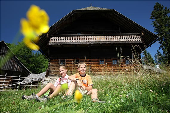 Blick auf das Geburthaus von Peter Rosegger und davor sitzen zwei Kinder in der Blumenwiese